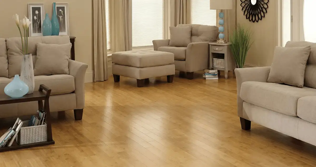 A living room with professionally installed bamboo flooring, beige sofas, a matching ottoman, a side table with a blue lamp, vases, framed pictures, and large windows with blinds and curtains in Erie, PA